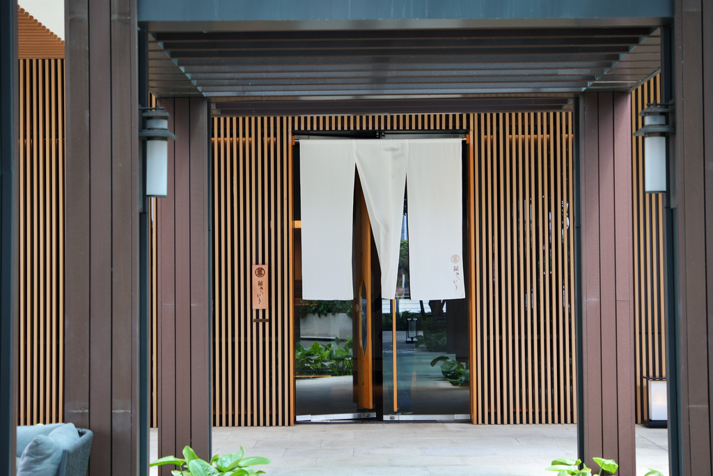 مدخل Sushi Saito في الجهة النهرية من Chao Phraya Estate، بستائر نورين بيضاء بين الشبكات الخشبية.