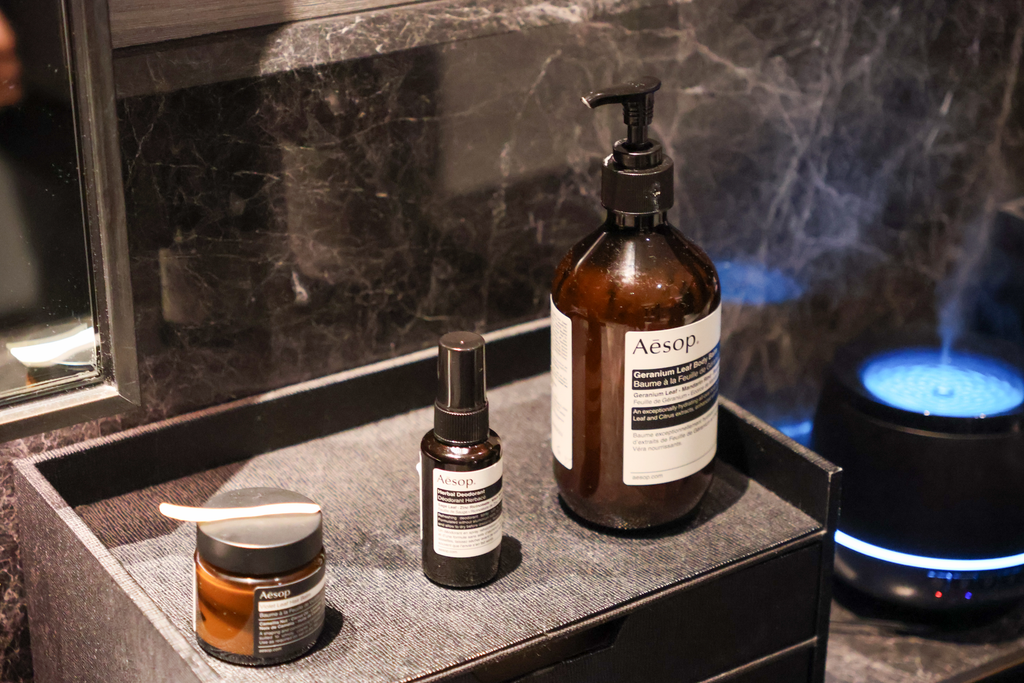 Violet Leaf Hair Balm وHerbal Deodorant وGeranium Leaf Body Balm من Aesop على صينية واحدة، جاهزة لتصفيف الشعر والعناية بالبشرة بعد الاستحمام.